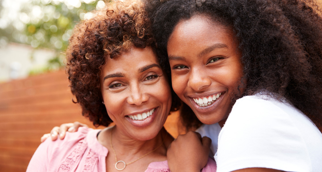 Middle aged black mum and teenage daughter embracing and smiling to camera